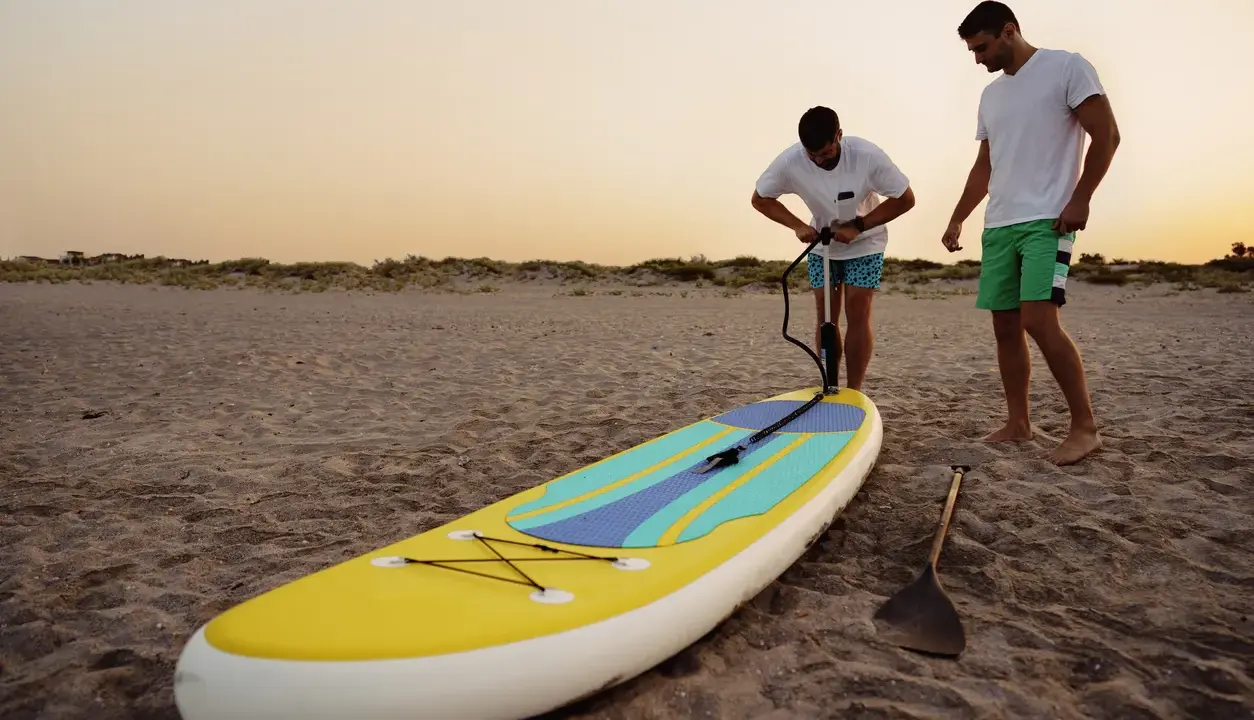 Zwei Männer bereiten ein SUP-Board am Strand vor. Einer pumpt es auf, fragt sich: "Sup, wieviel Bar?