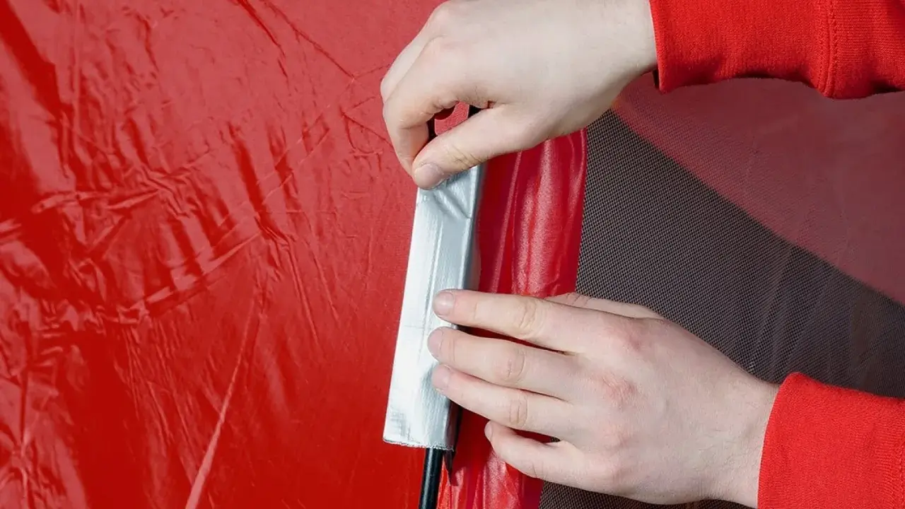 Hände flicken ein rotes Zelt mit silbernem Klebeband.