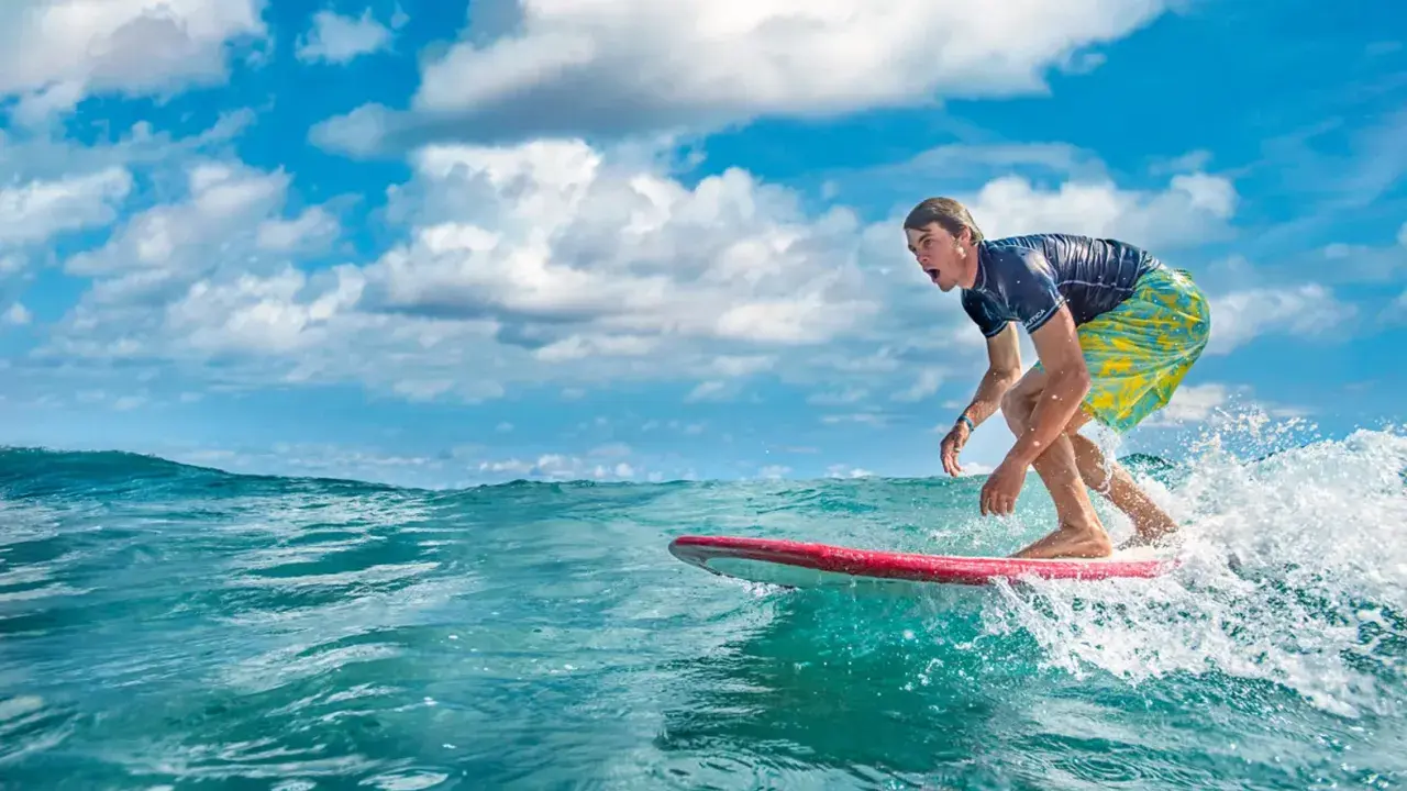 Ein Surfer reitet eine Welle vor der Küste der Dominikanischen Republik. Der Mann trägt ein blaues Shirt und bunte Badeshorts.