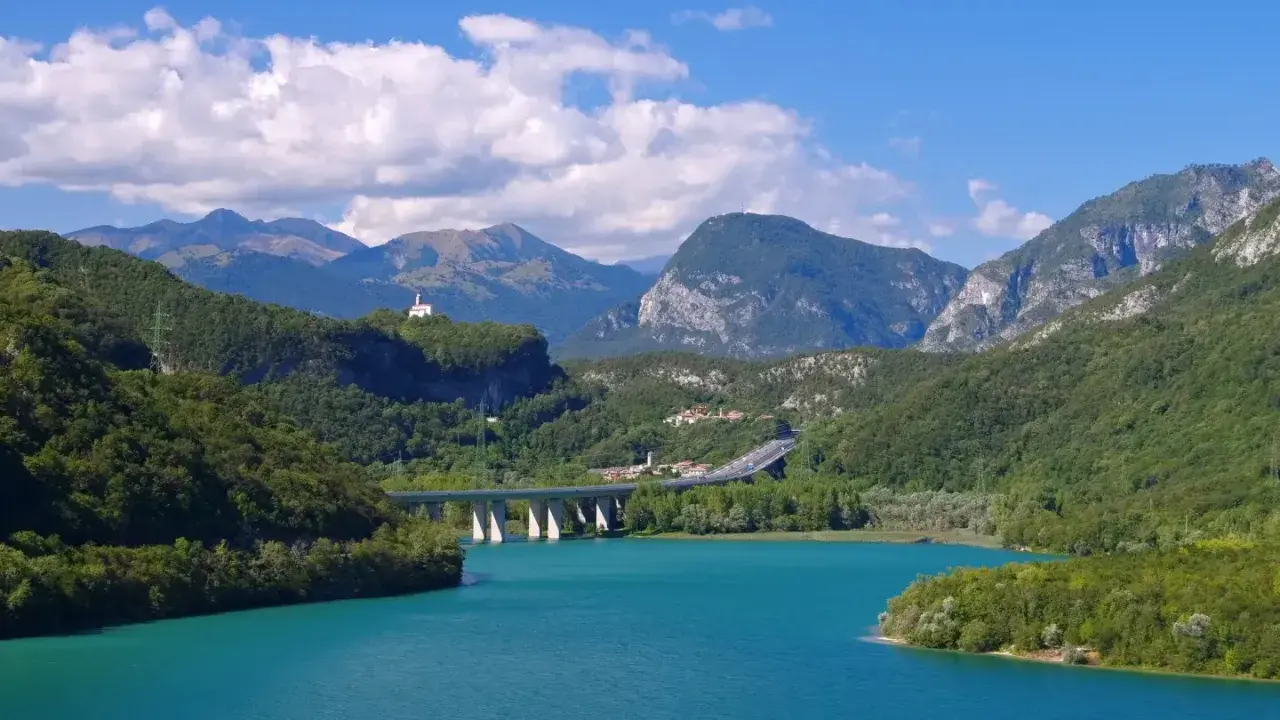 Türkises Wasser des Lago di Cavazzo umgeben von grünen Bergen. Eine Brücke überspannt den See, im Hintergrund eine kleine Kirche auf einem Hügel.