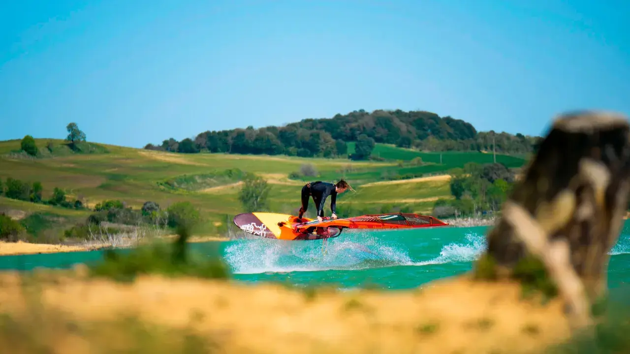 Windsurfer springt über das Wasser am Lac de Ganguise. Grüne Hügel und blauer Himmel im Hintergrund.