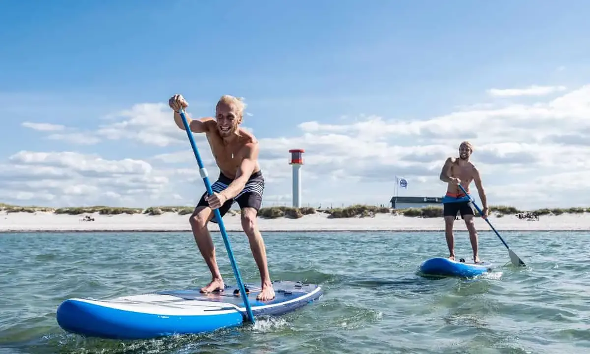 Zwei Männer genießen SUP auf der Nordsee. Sonnenschein, Strand und ein Leuchtturm im Hintergrund.