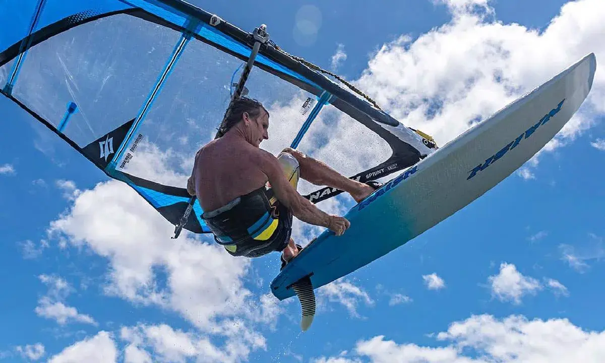 Ein Windsurfer mit einem Freeride Windsurfboard springt hoch in den blauen Himmel mit Wolken.