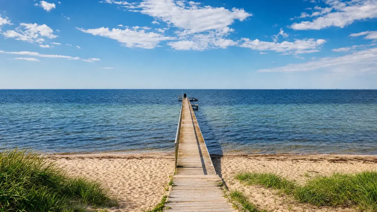 Holzsteg führt ins Meer am Ringkøbing Fjord. Eine Person steht am Ende des Stegs. Blauer Himmel mit Wolken.