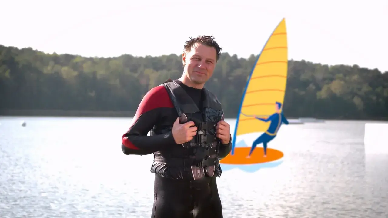 Mann in Neoprenanzug und Schwimmweste erklärt, wie funktioniert ein Neoprenanzug. Im Hintergrund eine stilisierte Windsurferin.