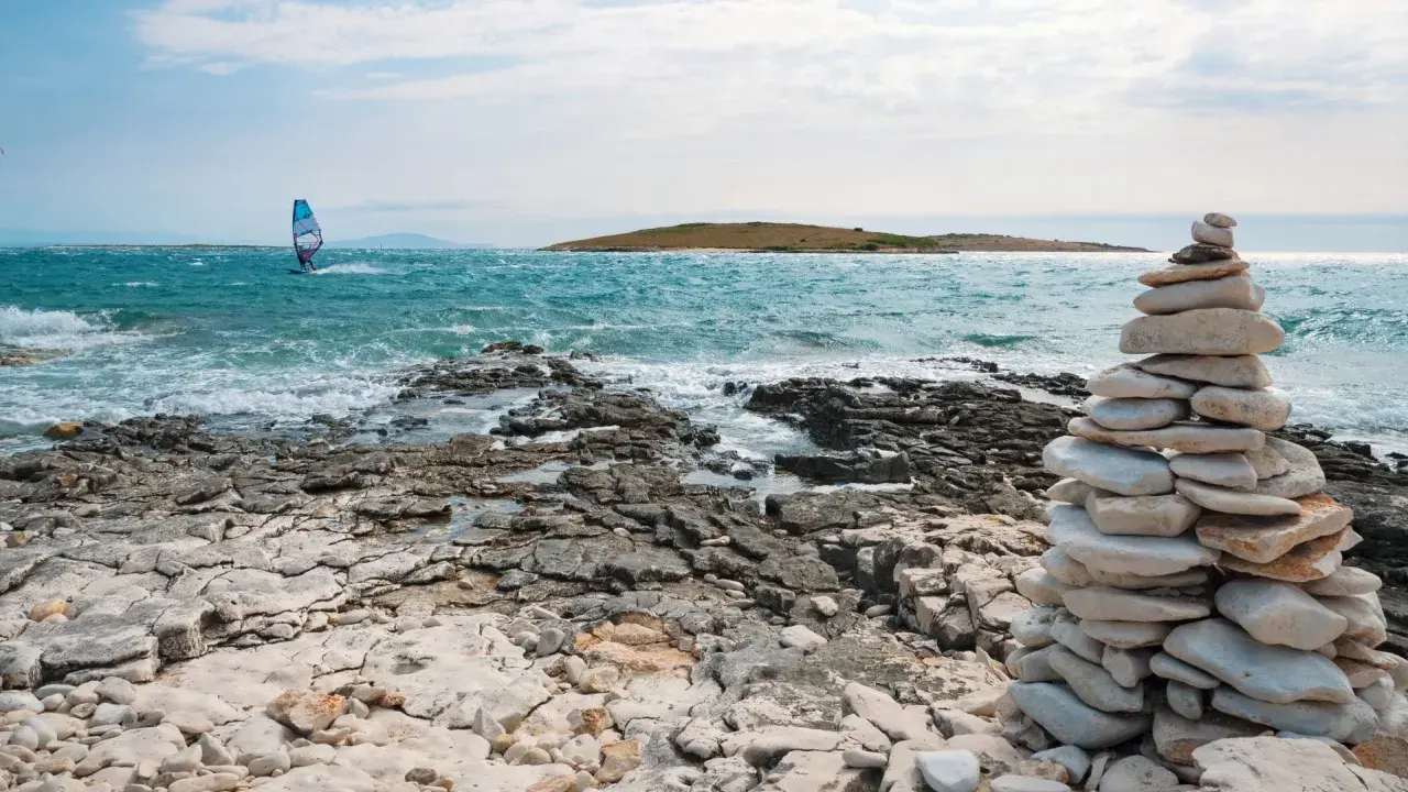 Ein Windsurfer gleitet über das türkisfarbene Meer vor Kroatien. Steinformationen säumen die felsige Küste.