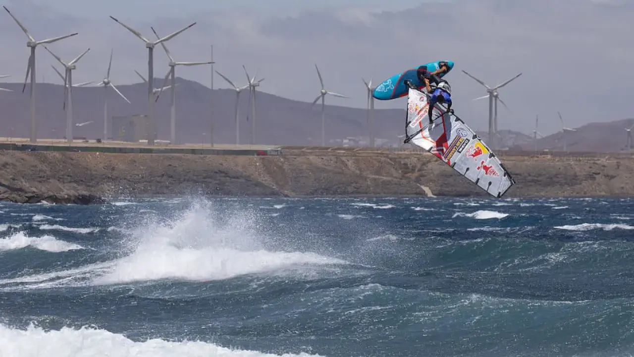Windsurfer Britt Dunkerbeck springt hoch über den Wellen, im Hintergrund Windräder.