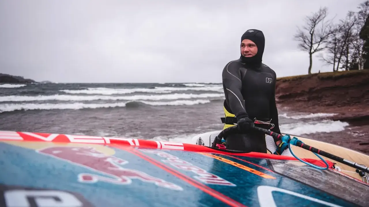 Mann in Neoprenanzug mit Windsurfbrett am Strand. Kaltes Wetter für Windsurfen in Deutschland.