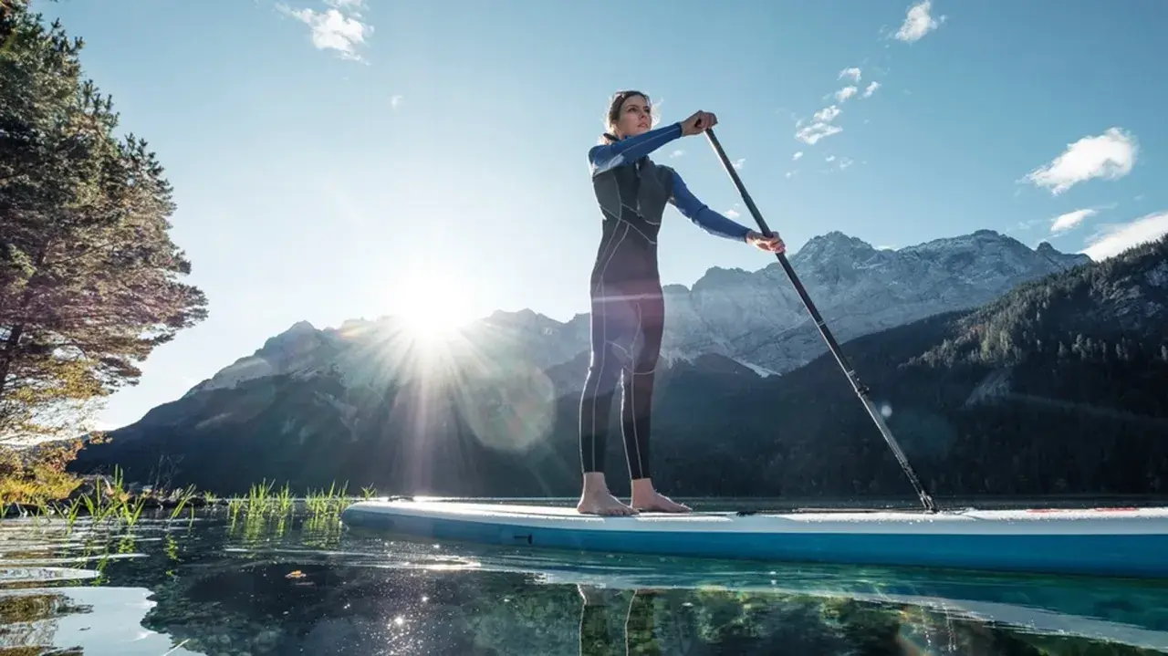 Eine Frau steht auf einem SUP-Board in Bayern, umgeben von Bergen und klarem Wasser.