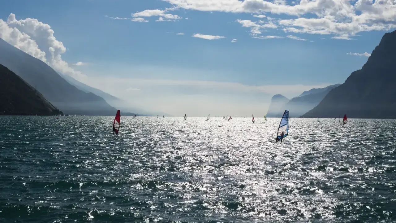 Windsurfer tummeln sich auf dem glitzernden Gardasee vor einer Bergkulisse.