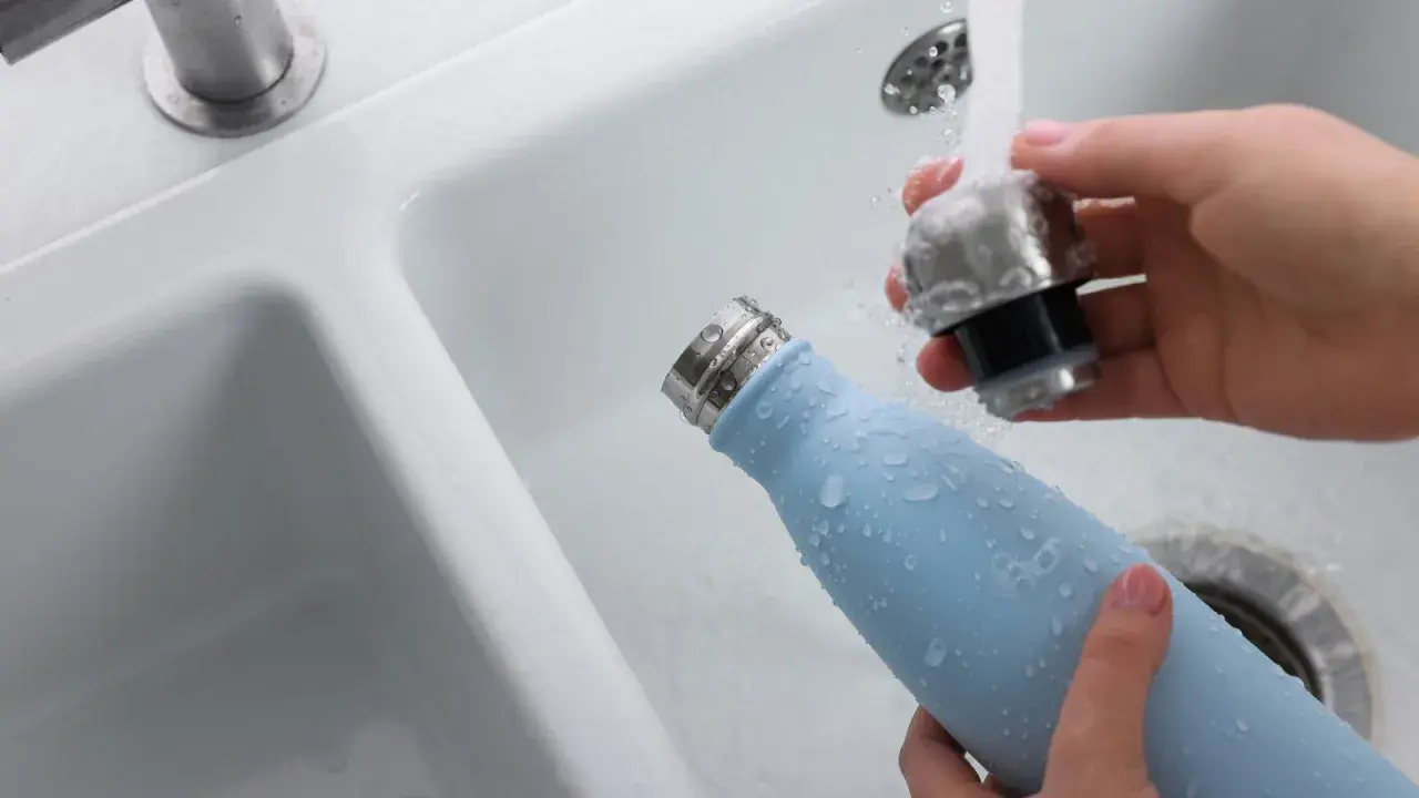 Hände reinigen eine hellblaue Trinkflasche unter fließendem Wasser im Waschbecken.