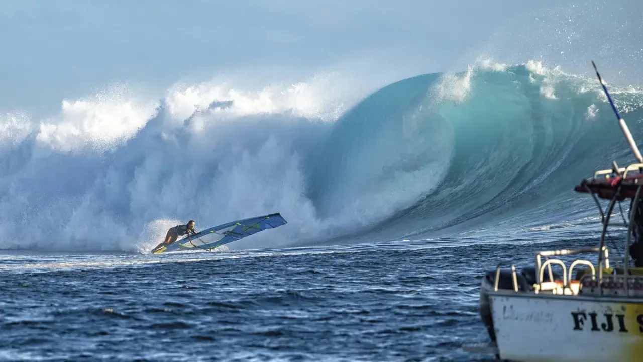 Sarah Hauser surft auf einer riesigen Welle, die sich hinter ihr bricht. Ein Boot mit der Aufschrift "Fiji S" liegt im Vordergrund.