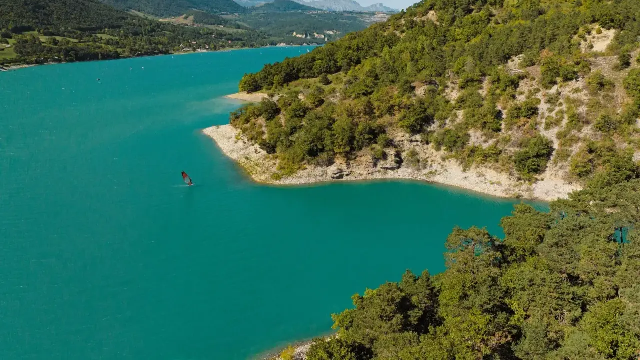 Türkisfarbener Lac de Monteynard mit einem Windsurfer, umgeben von bewaldeten Hügeln und Bergen.