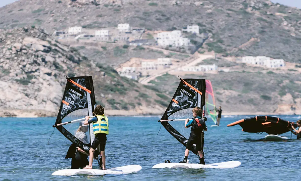 Kinder lernen naxos surfen. Zwei junge Windsurfer üben auf dem Meer, im Hintergrund weiße Häuser am Hang.