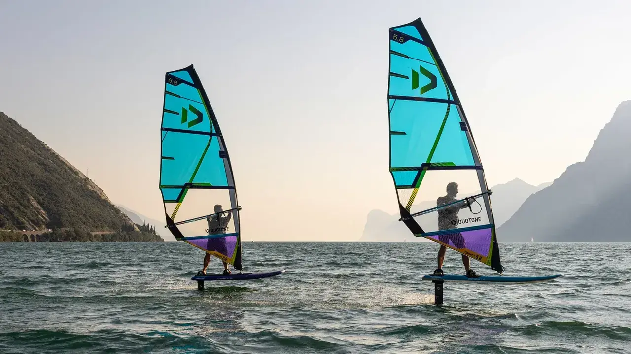 Zwei Personen beim Windsurfen in Italien. Die türkisfarbenen Segel sind im Gegenlicht zu sehen, im Hintergrund Berge.