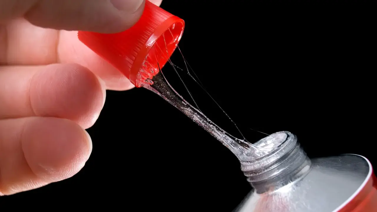 Ein Finger hält die rote Kappe eines Tubenklebers, aus dem zäher, glitzernder Kleber in Fäden heraustropft.