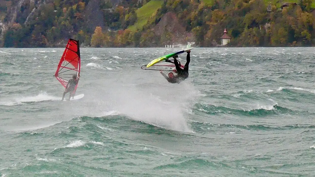 Windsurfer vollführt Sprung am Starnberger See. Ein weiterer Surfer mit rotem Segel im Hintergrund.