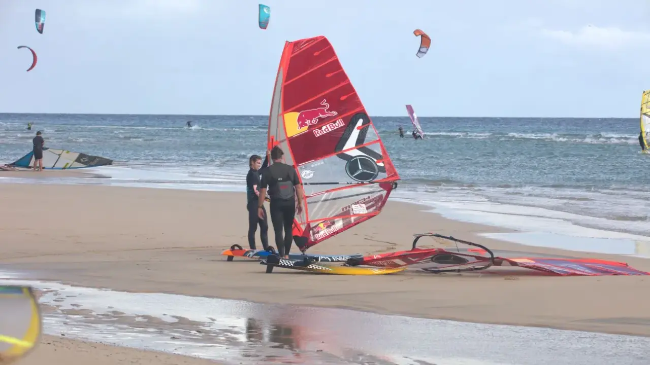 Windsurfer bereiten sich am Strand von Pozo Izquierdo, Gran Canaria, auf das Wasser vor. Mehrere bunte Segel und Boards liegen am Sand.