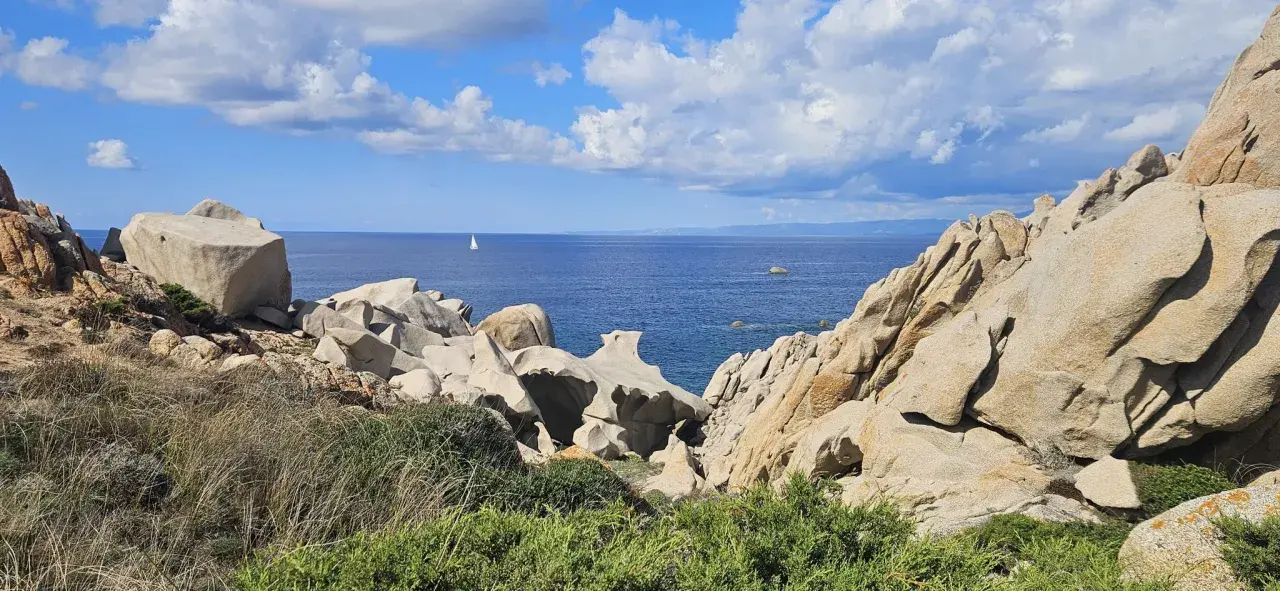Felsformationen am Capo Testa mit Blick auf das blaue Meer und einen einsamen Segler unter bewölktem Himmel.