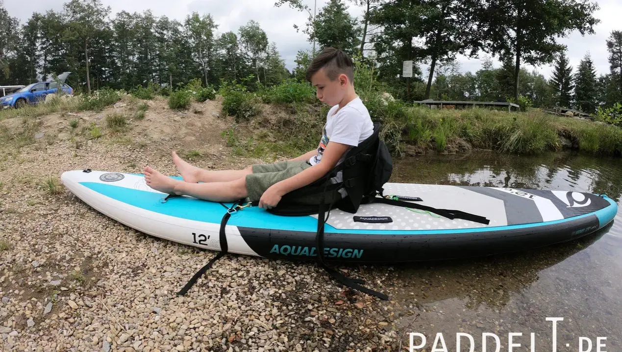 Junge sitzt auf SUP-Board, um den SUP Sitz zu befestigen. Das Board liegt am Ufer.
