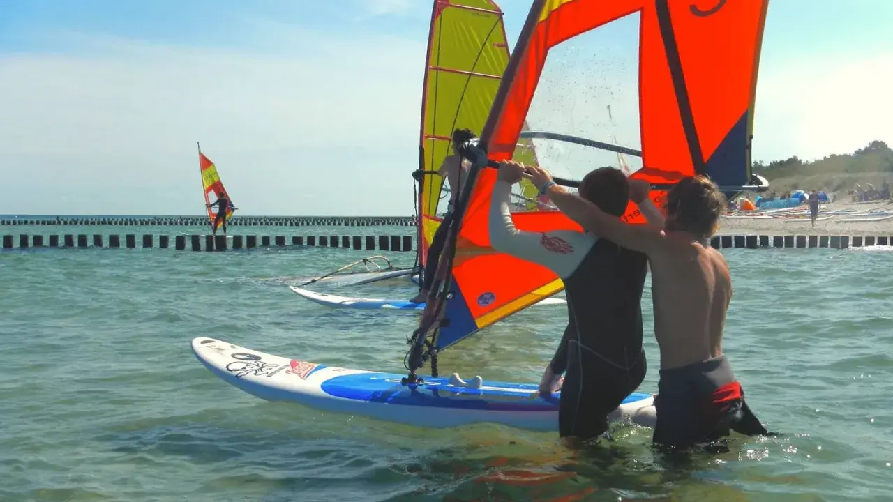 Zwei junge Männer bereiten sich auf das Windsurfen an der Ostsee vor. Ein weiterer Surfer ist in der Ferne zu sehen.