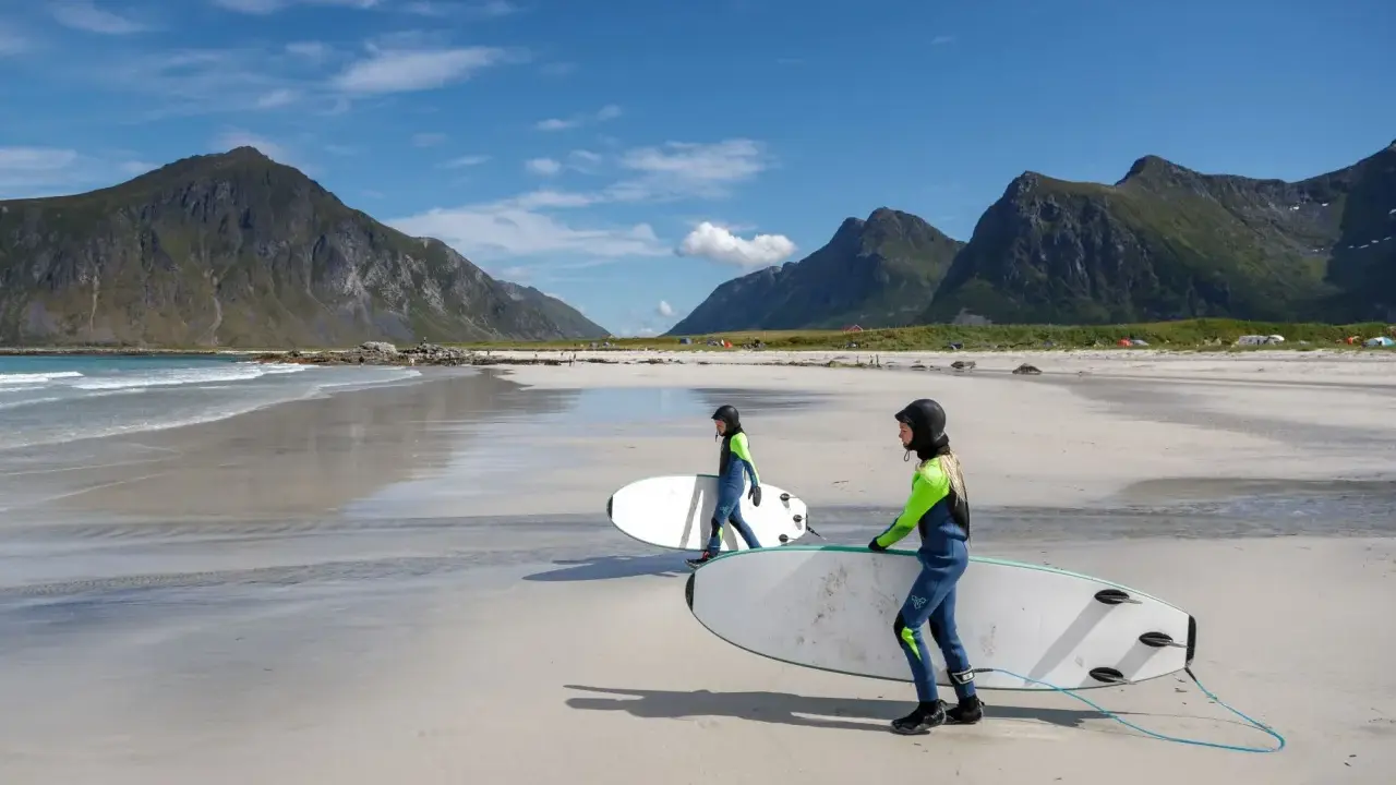Zwei Surferinnen mit Neoprenanzügen und Surfbrettern am Strand in Norwegen. Die Berge im Hintergrund sind grün und die Wellen sind sanft.