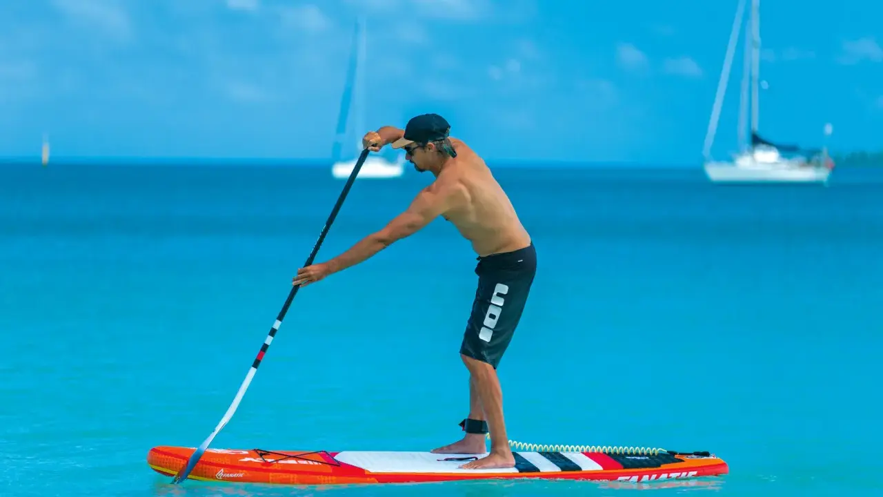 Mann stellt sein SUP Paddel ein, während er auf einem Board im türkisfarbenen Wasser steht. Segelboote sind im Hintergrund zu sehen.