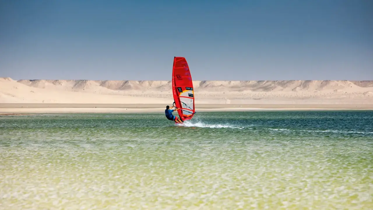 Windsurfer mit rotem Segel gleitet über das Wasser bei Dakhla, Marokko, vor einer Wüstenlandschaft.