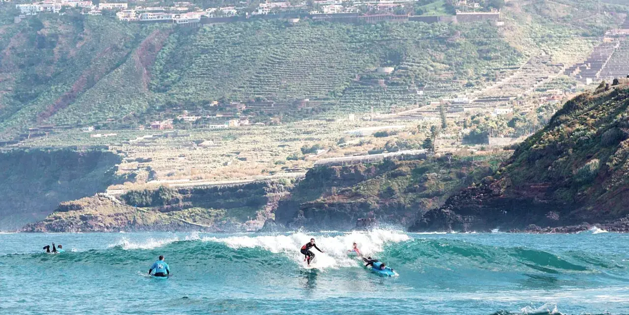 Surfer auf La Gomera reiten eine Welle, während im Hintergrund terrassierte Hänge und Häuser zu sehen sind.