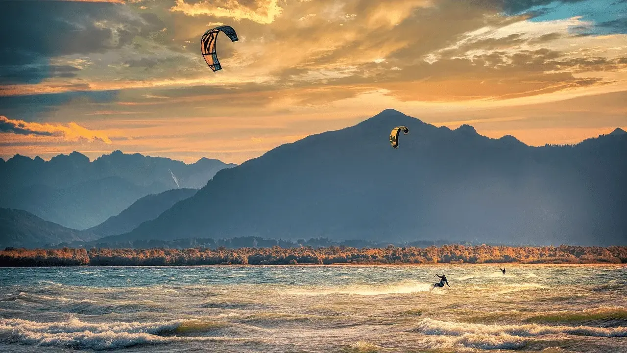 Kitesurfer gleiten über die Wellen am Chiemsee, während die Sonne hinter den Bergen untergeht. Ein traumhafter Abend für Windsurfen.
