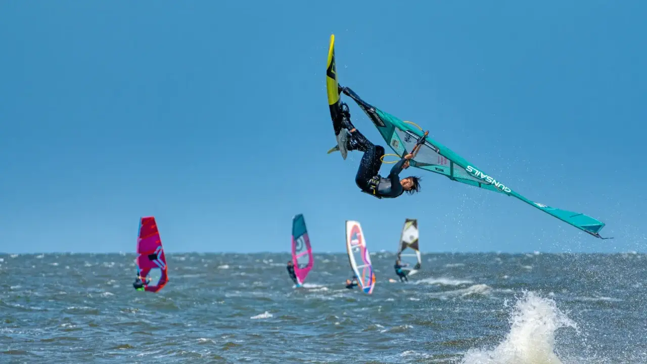 Windsurfer vollführt spektakulären Sprung über den Wellen. Mehrere andere Windsurfer sind auf dem Wasser zu sehen, was auf beliebte Windsurfing Spots hindeutet.