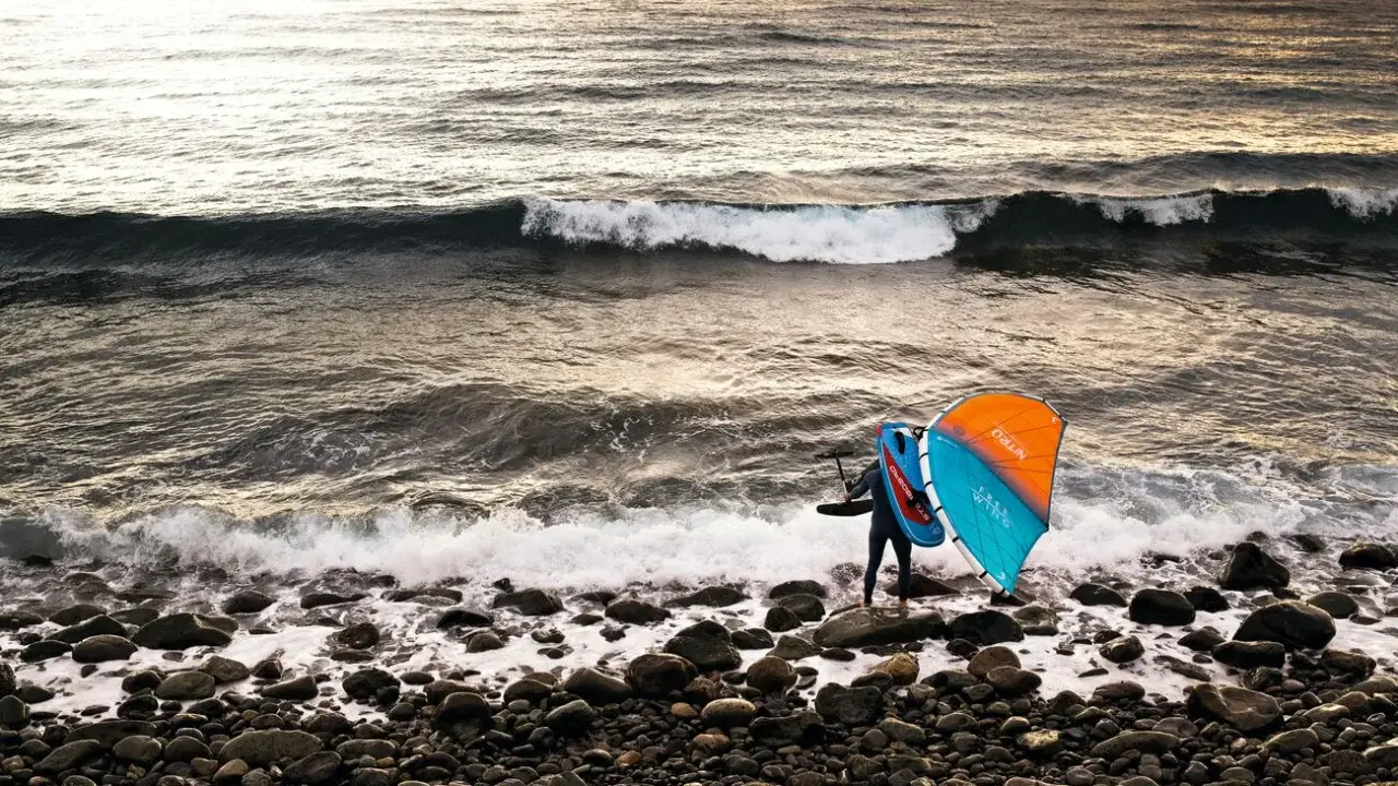 Ein Wingfoiler steht am steinigen Strand, bereit für den **shore break**. Die Sonne wirft ein warmes Licht auf die Wellen.