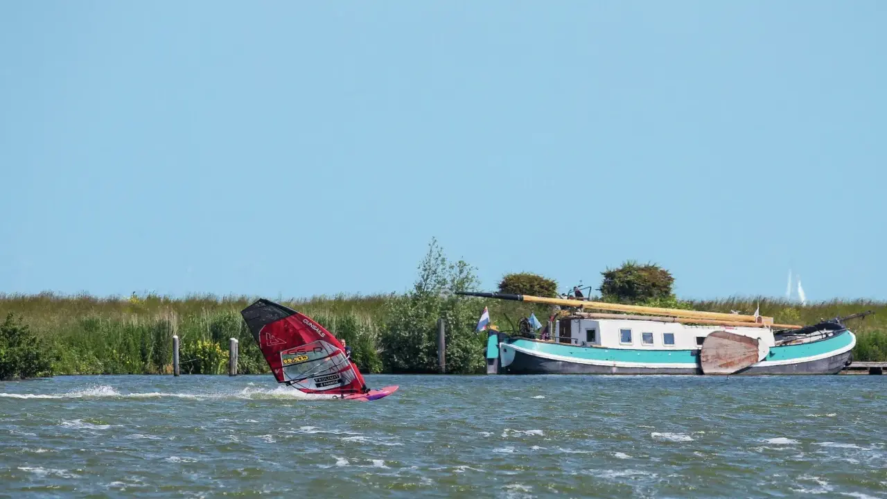 Windsurfen Holland: Ein Surfer gleitet über das Wasser, während ein Hausboot am Ufer liegt.