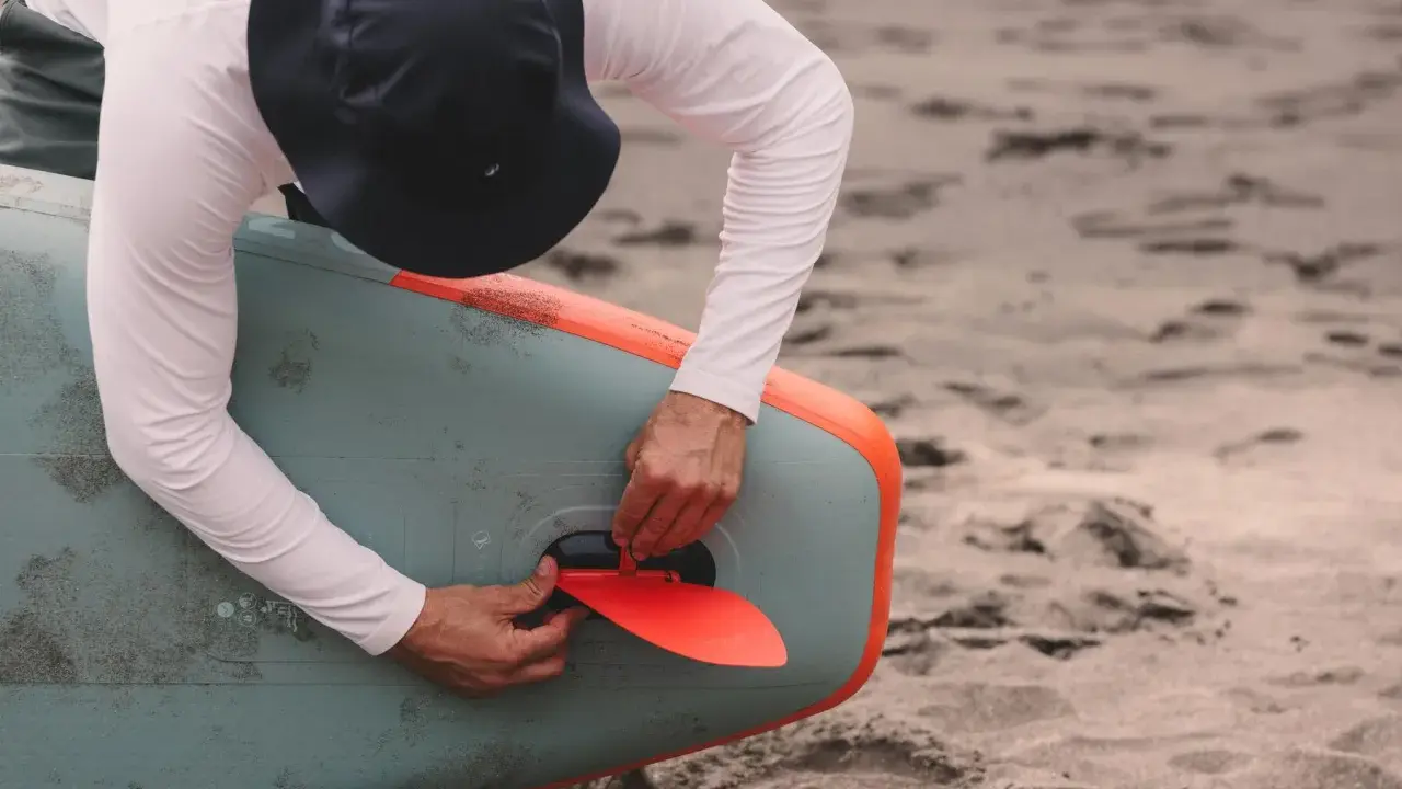 Mann mit Hut befestigt die rote Finne an einem aufblasbaren SUP-Board. Gute SUP-Pflege beginnt mit dem richtigen Zubehör.