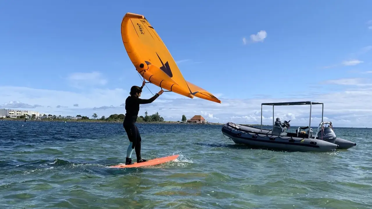 Person auf einem Board mit einem orangen Flügel, der über dem Wasser schwebt. Ein Schlauchboot liegt daneben. Foil surfen ohne Segel.