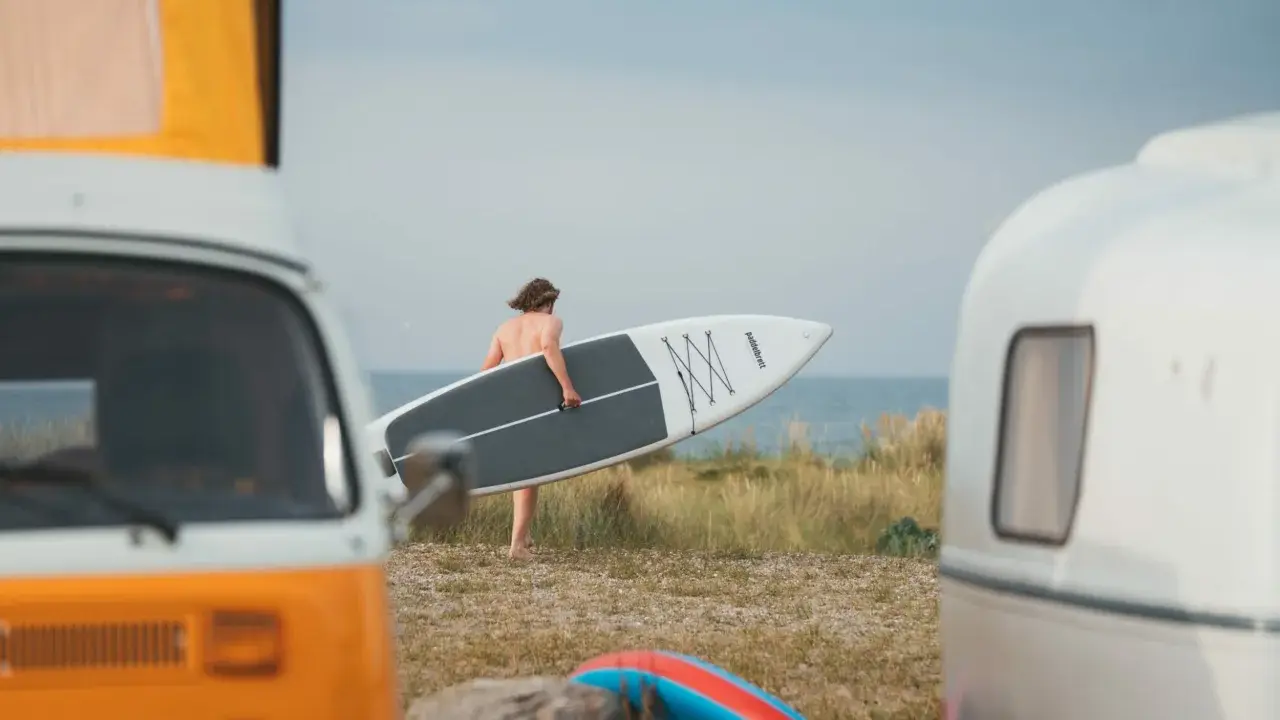Mann mit SUP-Board am Strand von Fehmarn, bereit für die Wellen. Wohnmobile im Vordergrund.