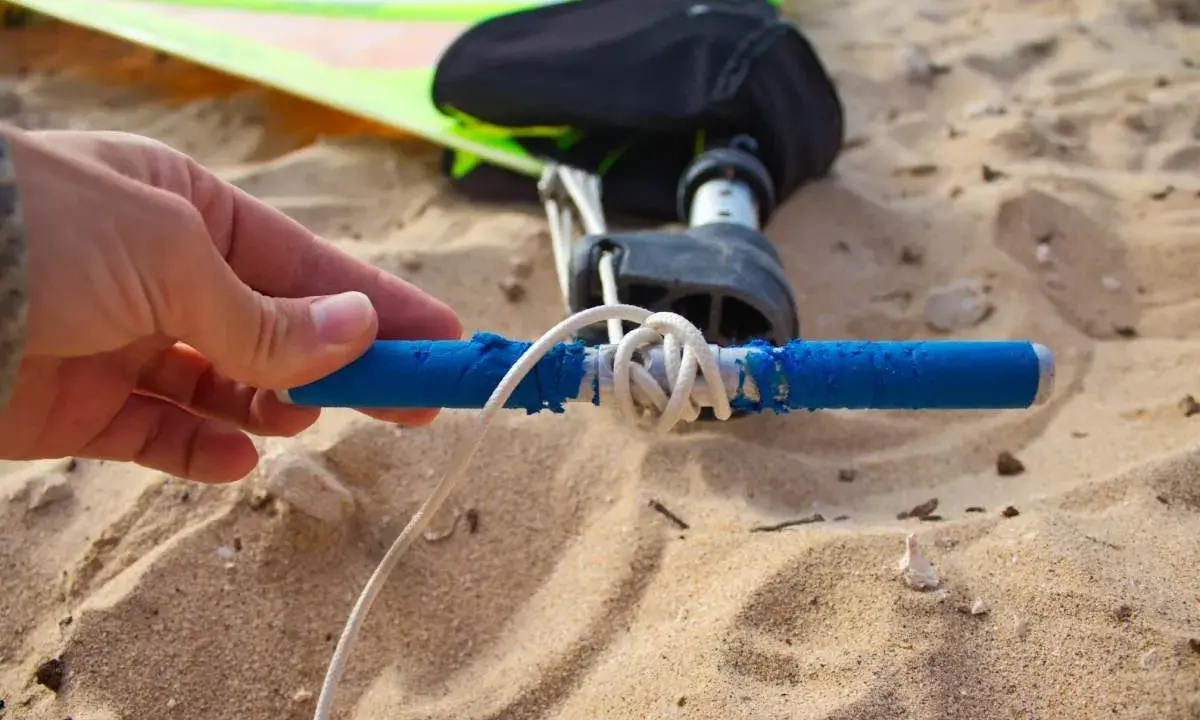 Hand hält blauen Griff, um das Segel zu spannen. Windsurf-Ausrüstung liegt im Sand.