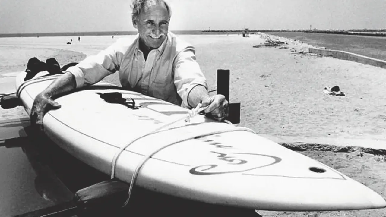 Jim Drake, ein Pionier des Surfens, lächelt stolz neben seinem Surfbrett am Strand.