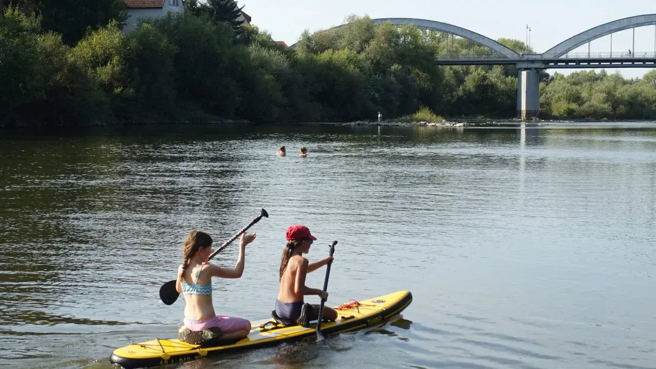 Zwei Mädchen genießen das Stand Up Paddling in Heidelberg. Im Hintergrund eine Brücke und Bäume.
