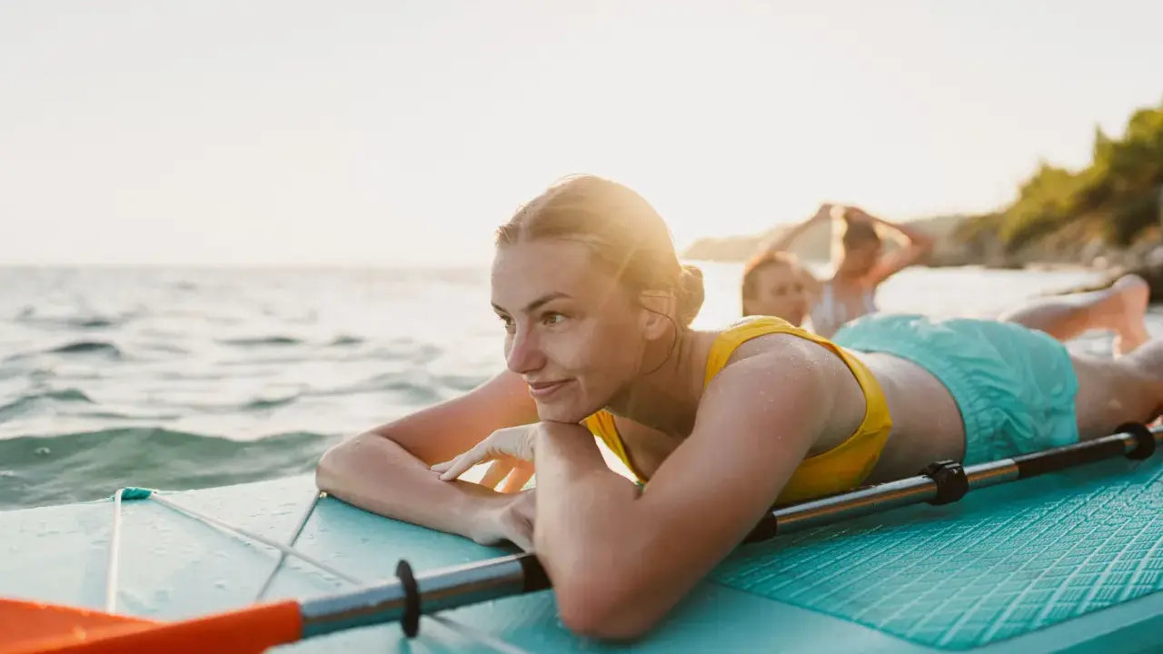 Junge Frau entspannt auf SUP-Board im Wasser. Ob Stand up paddling schwer ist, fragt sie sich lächelnd, während Freunde im Hintergrund planschen.
