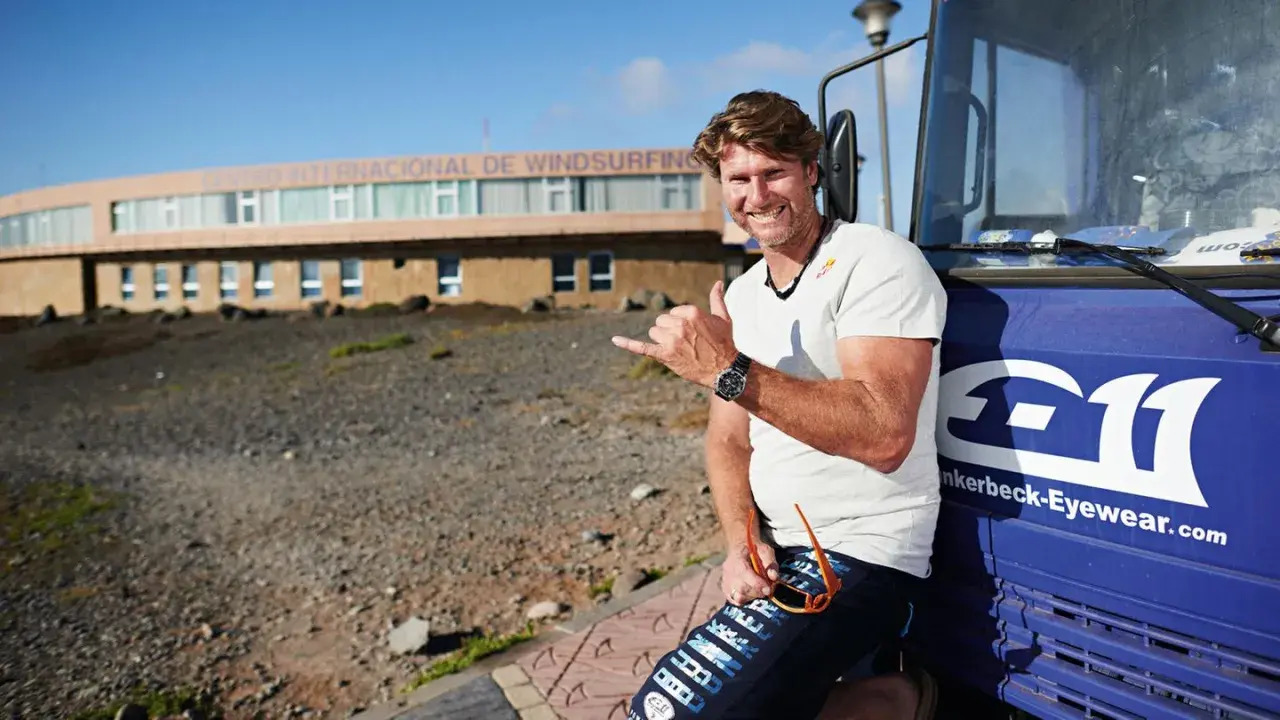 Bekannte deutsche Windsurfer: Ein Mann mit Sonnenbrille und Shaka-Geste lehnt an einem blauen Bus vor dem Centro Internacional de Windsurfing.