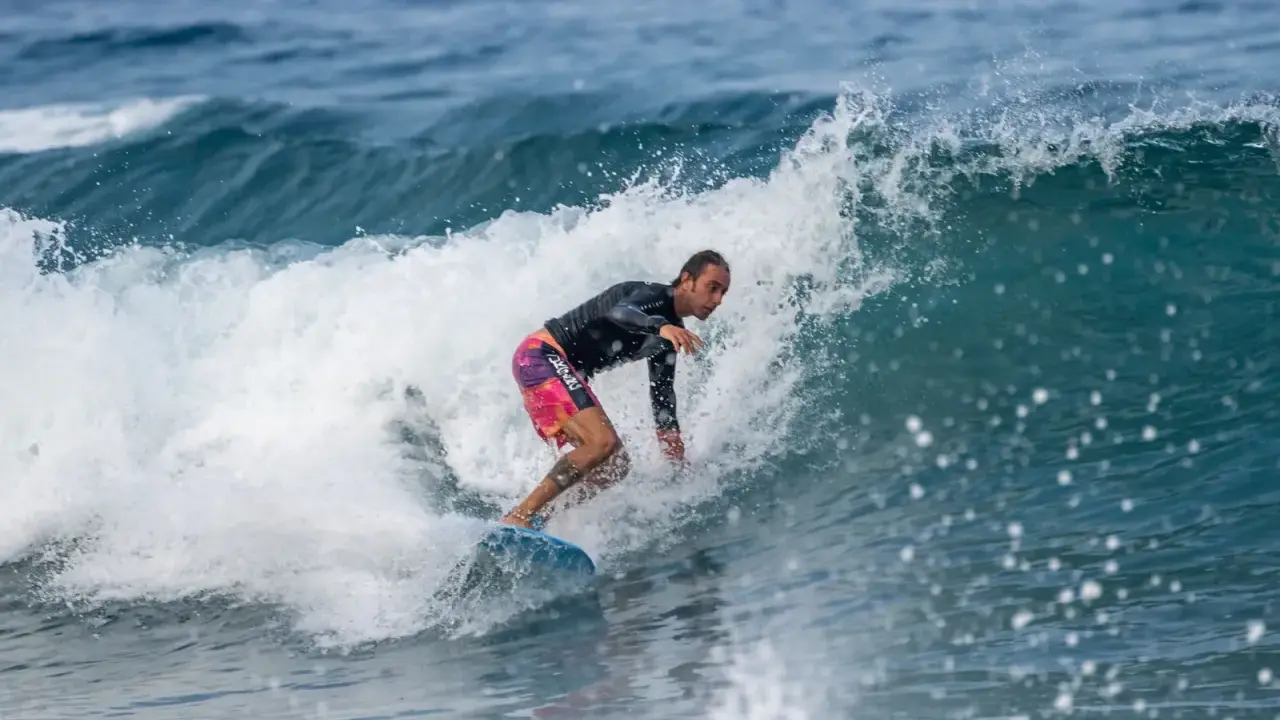 Ein Surfer reitet eine Welle vor Zypern. Er trägt ein schwarzes Rashguard und bunte Boardshorts.