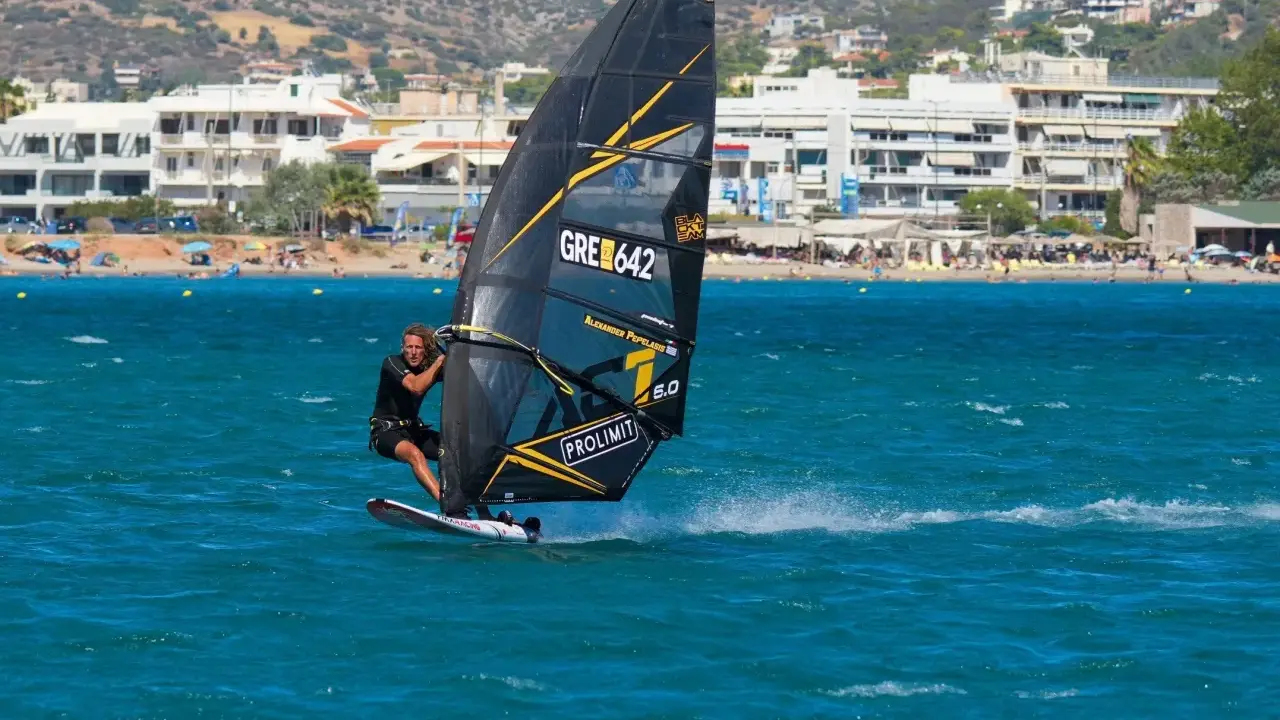 Ein Mann beim Windsurfen in Griechenland, mit Blick auf eine belebte Küste und Berge im Hintergrund.