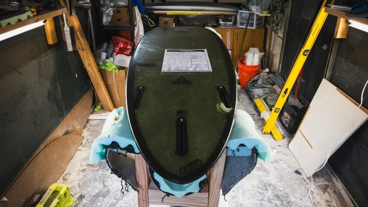 Surfboard liegt auf Ständer, bereit für die **Surfboard Reparatur**. Werkzeuge und Materialien im Hintergrund.