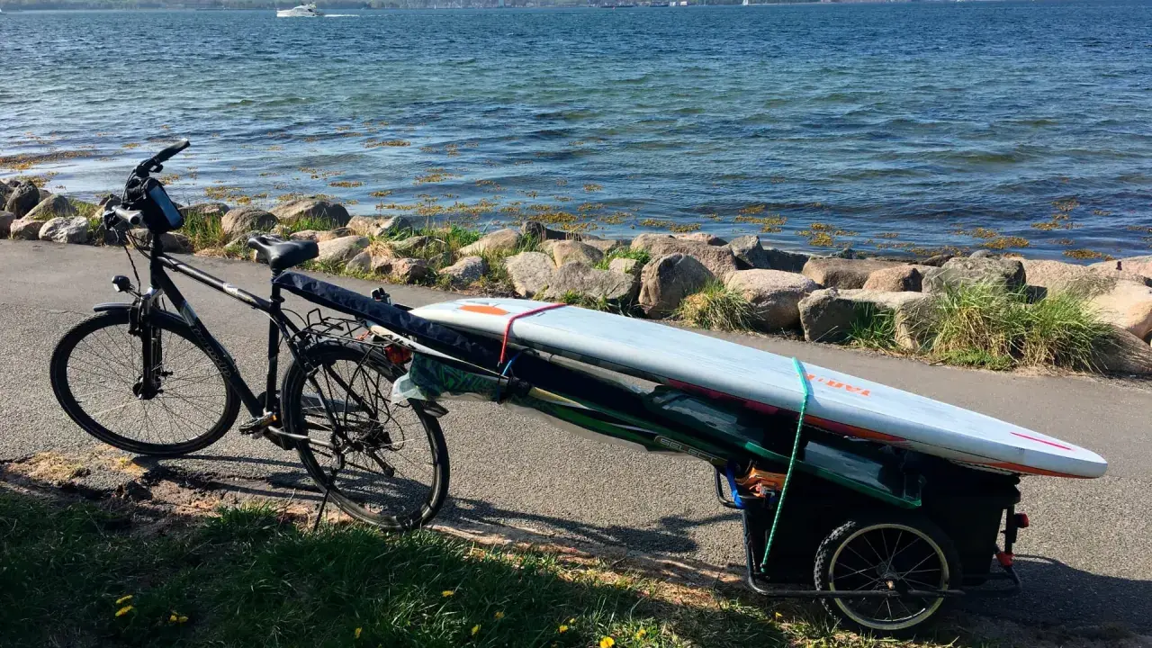Fahrradanhänger mit Surfbrett und Paddel am Seeufer, bereit für ein Abenteuer auf dem Wasser.