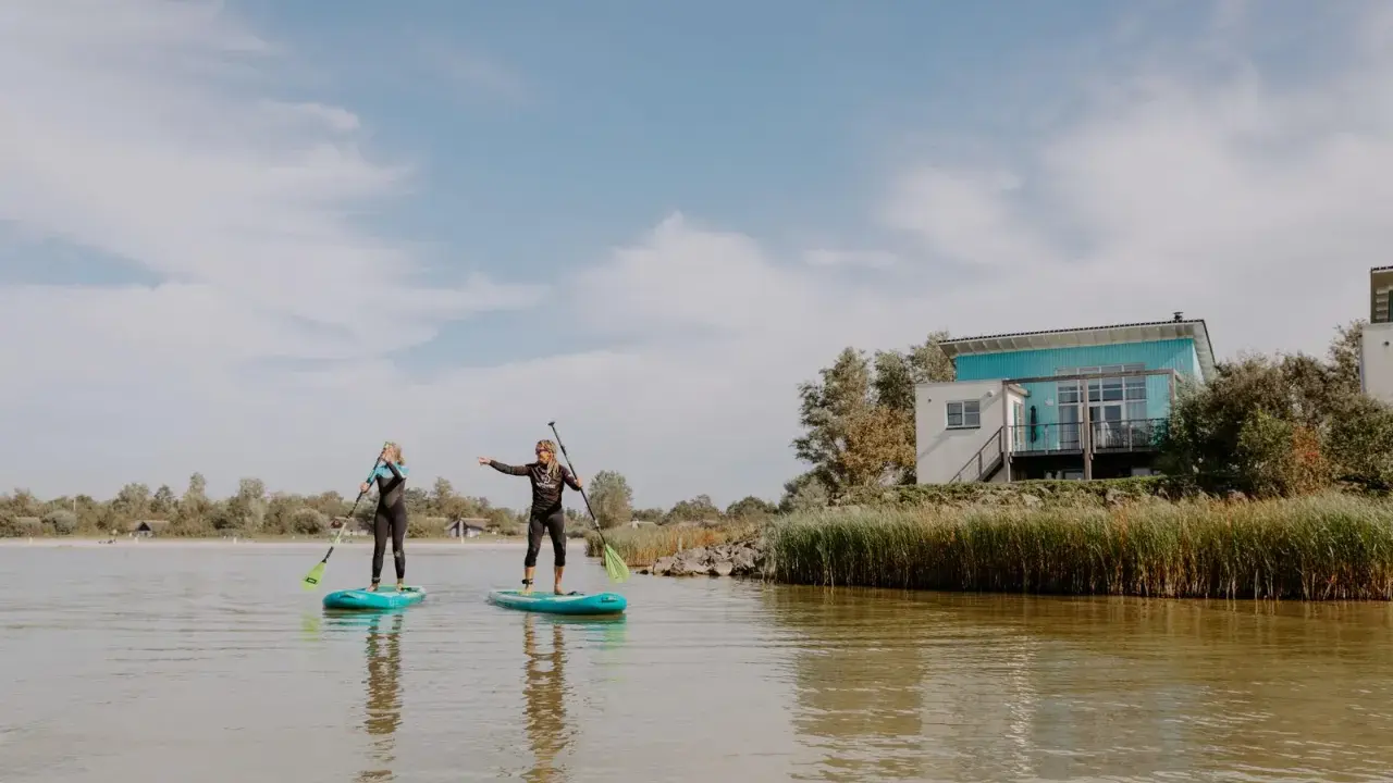 Zwei Frauen auf SUP-Boards am Makkum Strand, mit einem modernen Haus im Hintergrund.