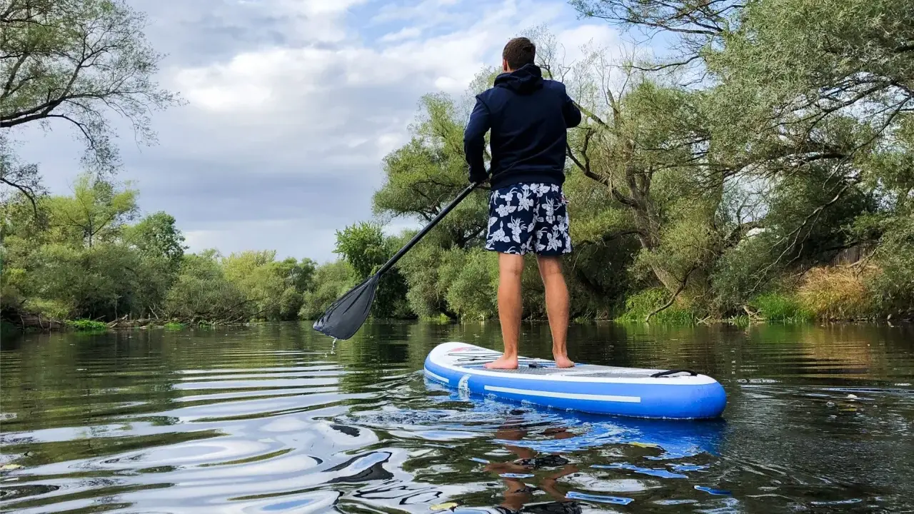 Mann auf SUP-Board auf ruhigem Fluss, umgeben von grünen Bäumen. Perfekt, um zu sehen, wo man stand up paddeln kann.