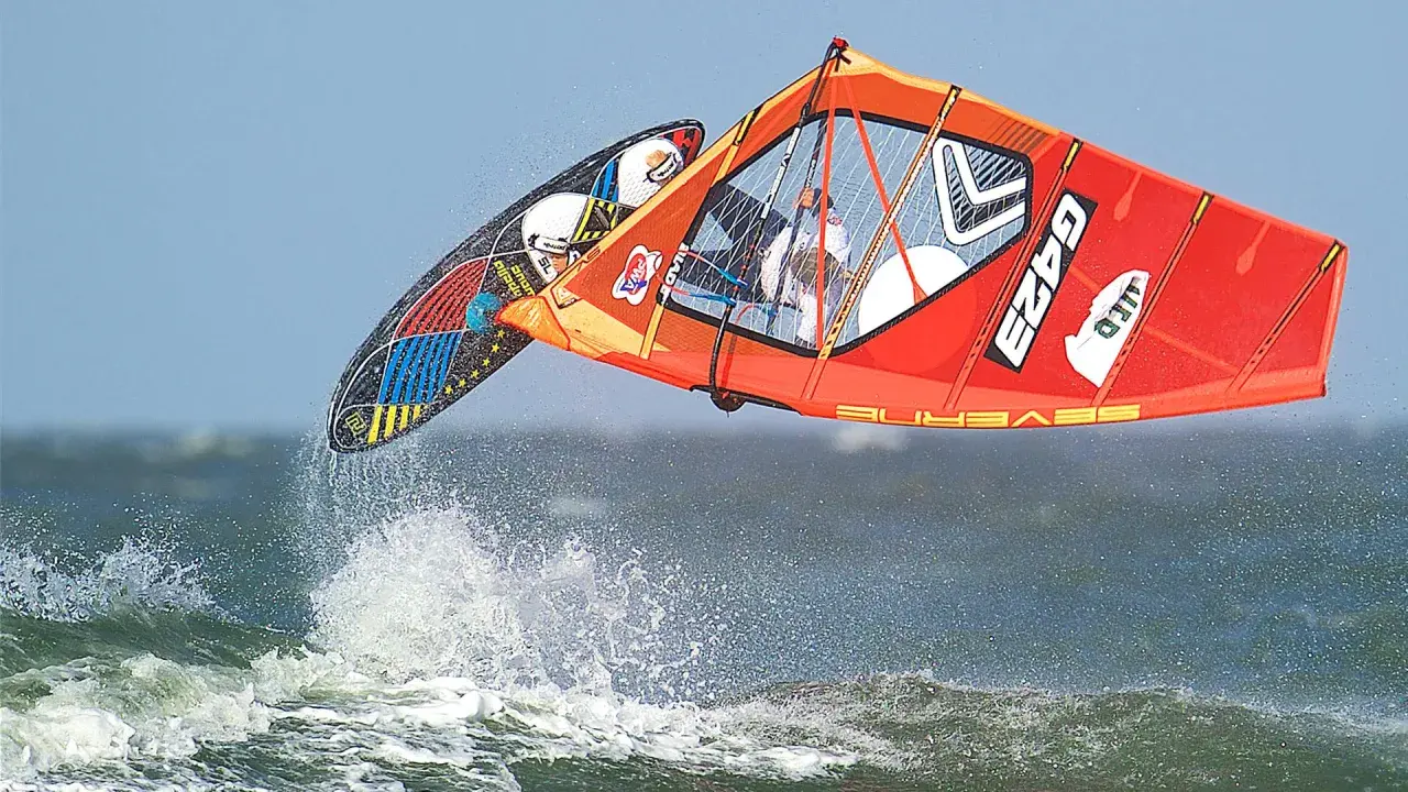 Lina Erpenstein fliegt mit ihrem Windsurf-Equipment über die Wellen. Das orangefarbene Segel und das bunte Board sind in der Luft.