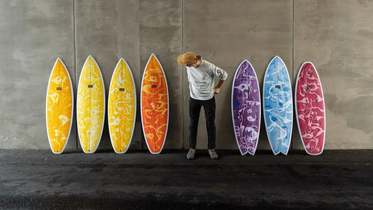 Mann mit Hut steht vor bunten Surfbrettern, bereit für das Wellenreiten.