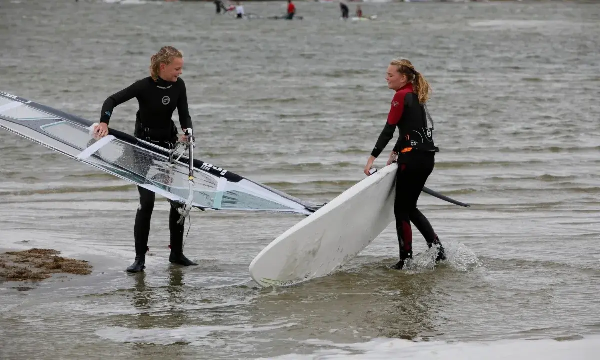 Zwei Mädchen bereiten sich auf das Windsurfen am IJsselmeer vor, tragen Neoprenanzüge und halten ihre Ausrüstung.